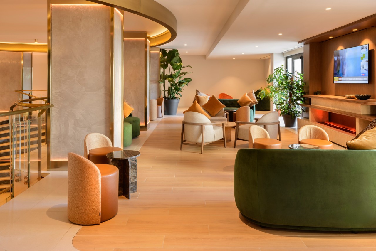Lobby de hotel moderno y elegante con suelo de madera, sillones de diseño en tonos verdes y terracota, iluminación cálida, plantas decorativas y zona de televisión con chimenea